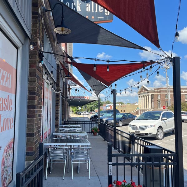 Outdoor patio of Back Alley Pizza with tables, chairs, red and black awnings, and string lights.