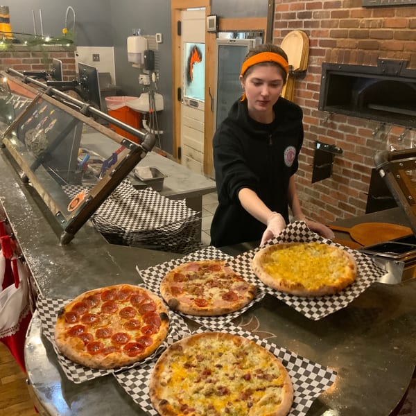 A Back Alley Pizza employee presents four freshly baked pizzas on checkered paper, with a brick oven in the background.