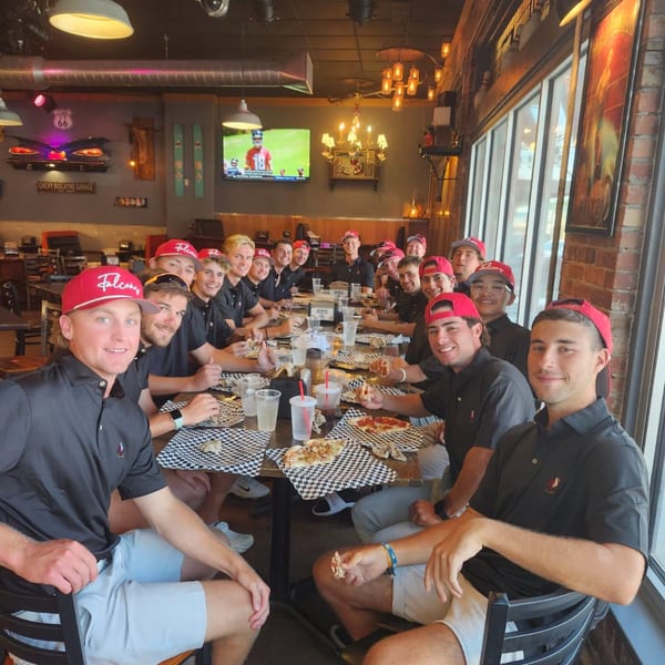 A large group of young men in red hats and black shirts eating pizza at Back Alley Pizza.